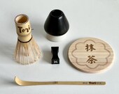 Personalized matcha tea set featuring engraved bamboo whisk and scoop with rhinestones, ceramic black whisk holder, scoop rest, and double-sided engraved wooden coaster. One side of the coaster shows Japanese matcha characters, the other features mat thumbnail 1