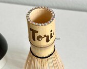 Personalized matcha tea set featuring engraved bamboo whisk and scoop with rhinestones, ceramic black whisk holder, scoop rest, and double-sided engraved wooden coaster. One side of the coaster shows Japanese matcha characters, the other features mat thumbnail 8