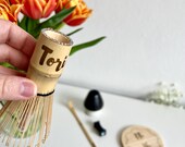 Personalized matcha tea set featuring engraved bamboo whisk and scoop with rhinestones, ceramic black whisk holder, scoop rest, and double-sided engraved wooden coaster. One side of the coaster shows Japanese matcha characters, the other features mat thumbnail 2